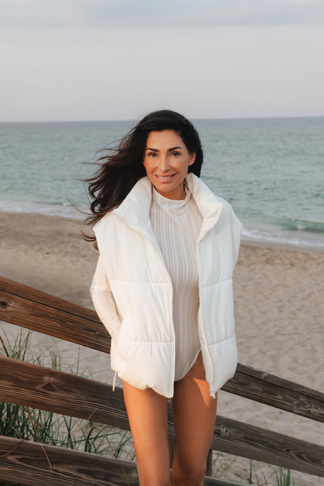 Dara T in a white puffer jacket standing on a wooden bench by the beach