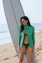 Woman in green bikini and green moto jacket standing on a beach with a surfboard.