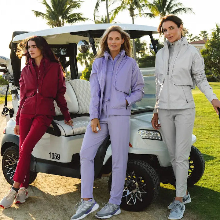Three women in golf attire standing next to a golf cart on a golf course.
