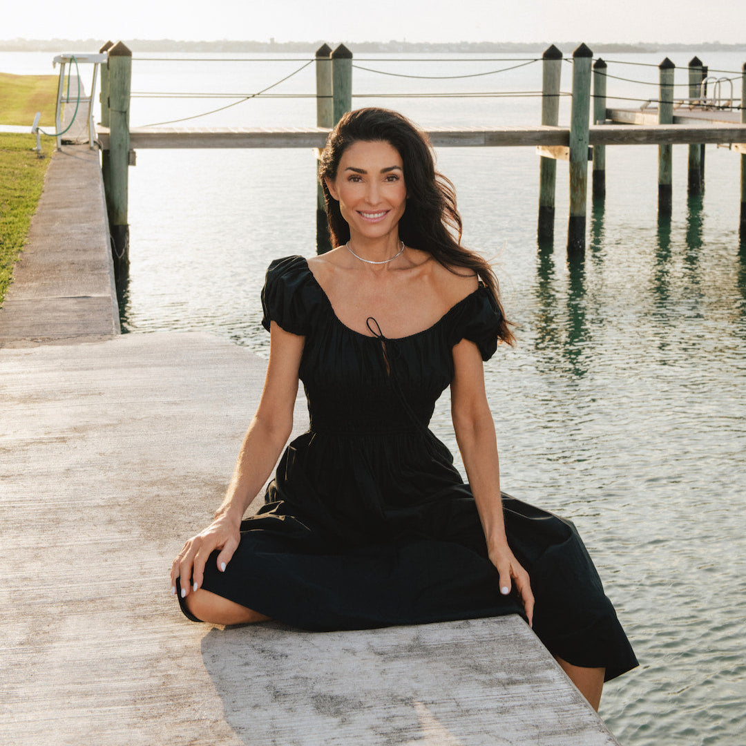Dara in a black dress sitting on a wooden dock by a body of water.