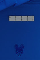 Color:Blu Close-up of a royal blue fabric garment, likely a shirt or jacket, featuring a horizontal row of five rectangular silver reflective tee holder and a small embroidered dog head motif below.