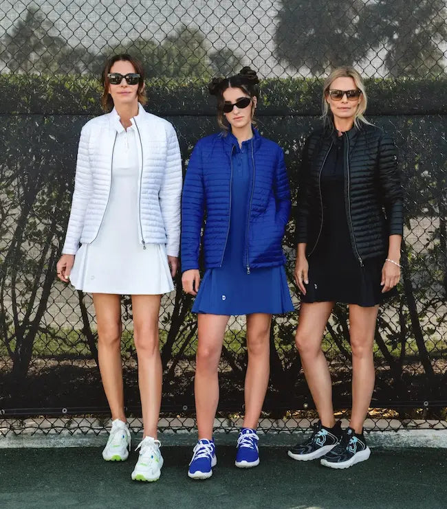 Three women standing on a tennis court wearing stylish puffer jackets.